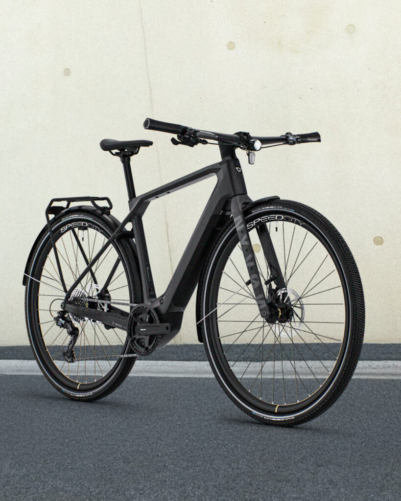 Foto eines mattschwarzen E-Bikes für Pendler mit Gepäckträger und grobstolligen Reifen, das vor einer hellen Betonwand steht.